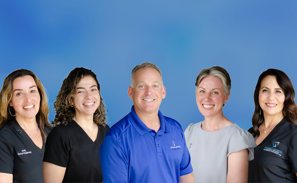 The image shows a group of smiling individuals posing together, with a man standing in the center and others on either side, all dressed in professional attire  they appear to be part of an organization or team.