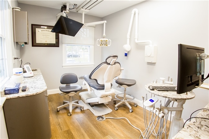 The image shows a dental office interior with a modern chair, equipment, and a desk with a computer monitor and keyboard, all set against a wall with a framed diploma and a window.