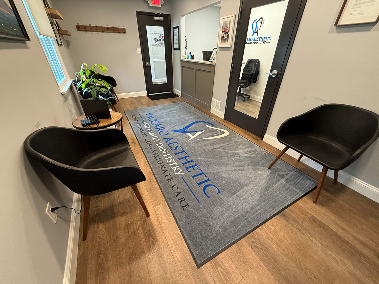 The image shows an office interior with a reception area featuring a rug with text, a logo on the wall, a desk, chairs, and decorative elements.