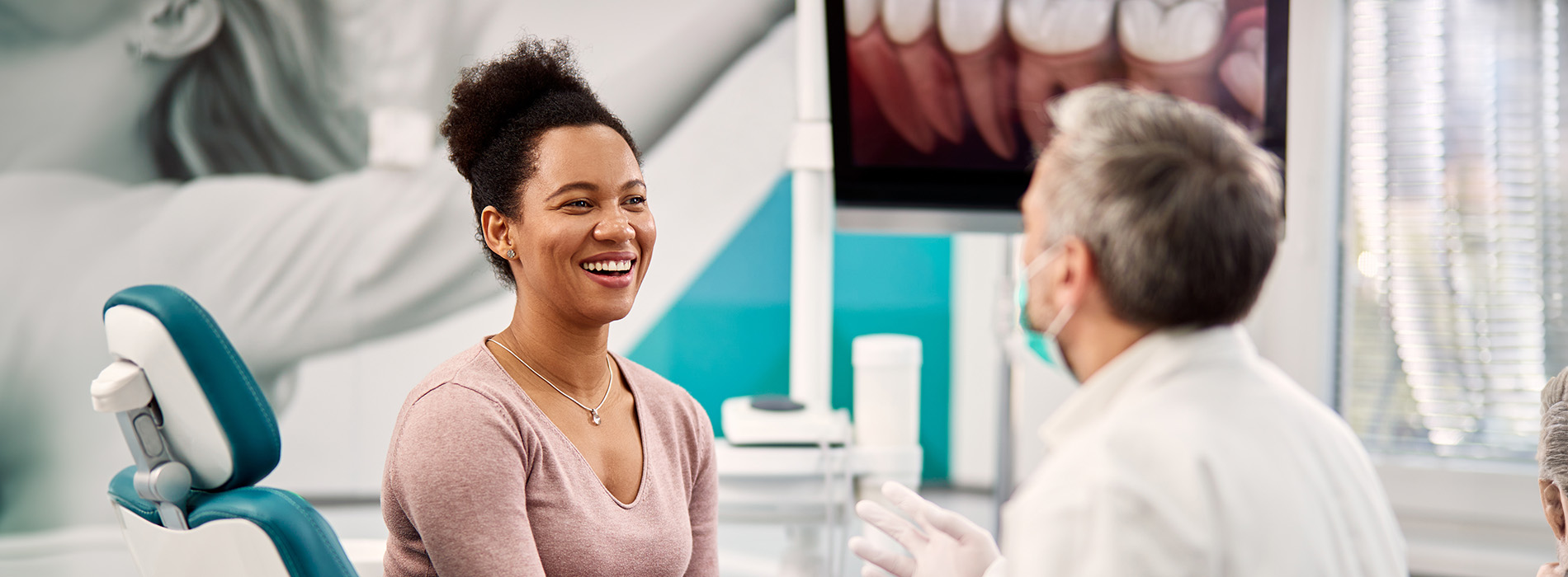 The image shows two people sitting in a dental office, with one person smiling at the camera and another seated next to them.