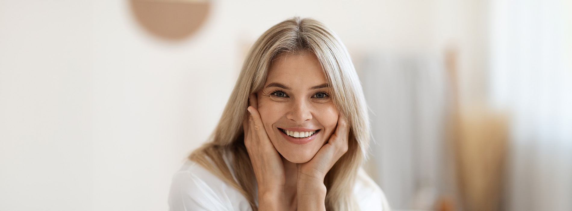 The image is a photograph of a woman with light skin, smiling at the camera. She appears to be in her late twenties or early thirties and has long hair. Her eyes are looking directly at the camera, and she is holding up her index finger near her mouth as if she s making a point or emphasizing something. The background is plain and light-colored, which suggests that this could be a stock photo used for various purposes such as advertising, personal branding, or lifestyle content.