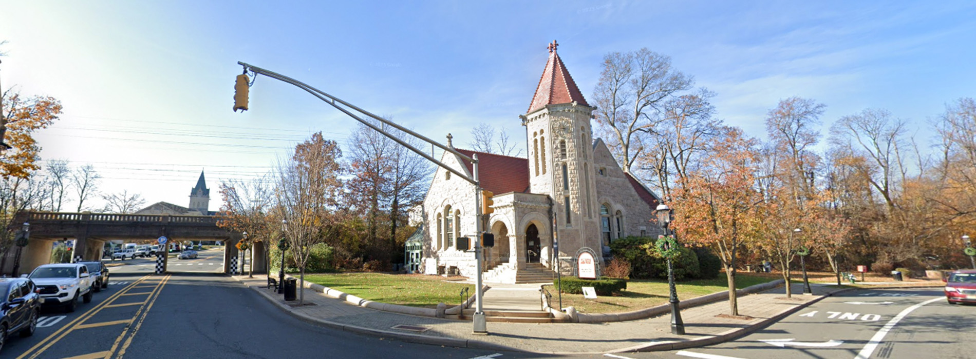 The image depicts a church with a prominent steeple, set against a blue sky, with trees showing autumn colors, and appears to be taken from a moving vehicle as indicated by reflections and distortions.