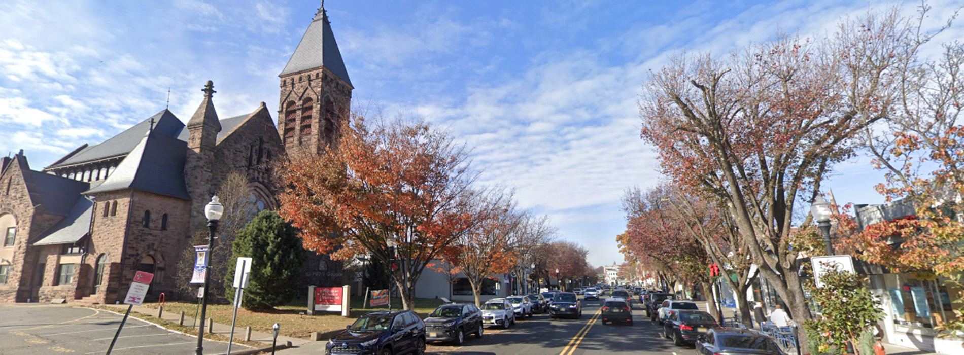The image depicts a street view of a town with a large church at the end, showcasing autumn foliage on trees along the roadside.