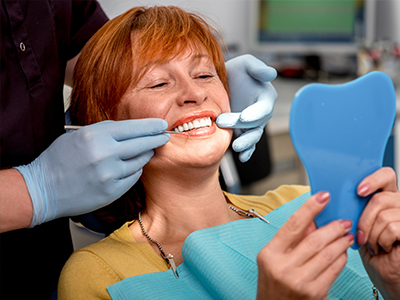 The image shows a woman sitting in a dental chair with her mouth open, receiving dental care from a professional who appears to be examining her teeth. She has red hair and is holding a blue toothbrush up to the camera, while looking directly at it.