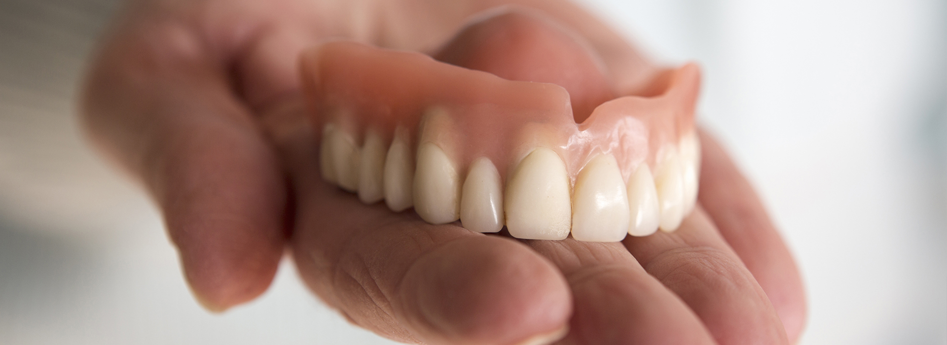 The image shows a person holding a set of dentures with their hand, displaying the artificial teeth against a blurred background.