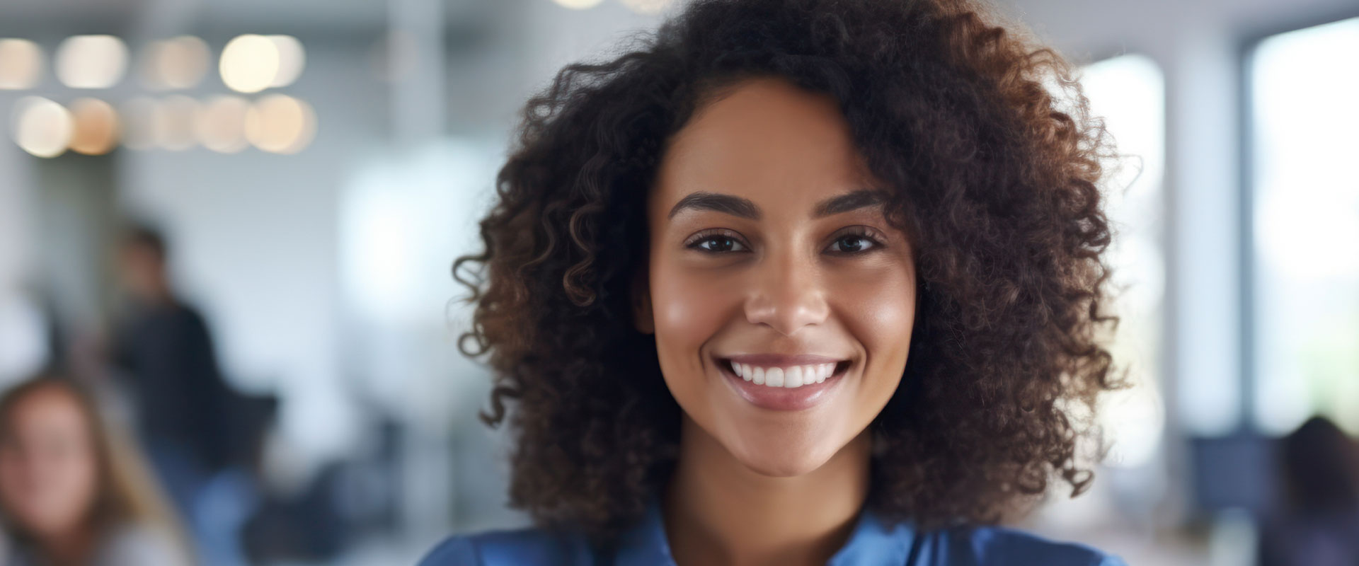 The image features a woman with curly hair smiling at the camera, wearing a professional attire with a blue top and a white background behind her.