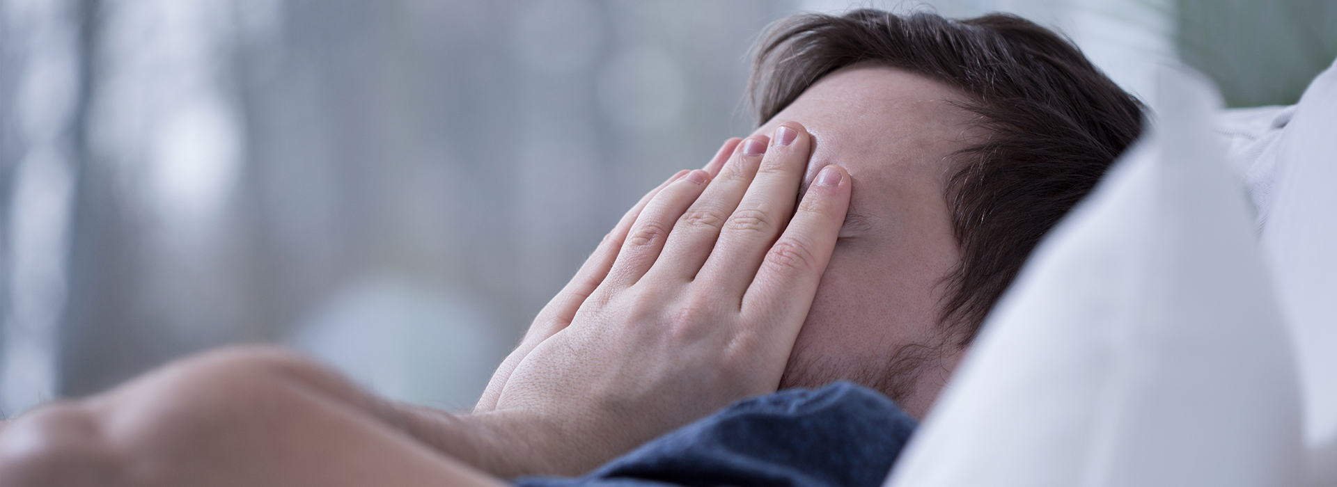 A man lying down with his hands over his face, looking distressed or surprised, set against a blurred background.