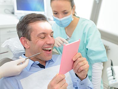A man is sitting in front of a pink card being held by a dental professional, smiling at the camera while holding a phone with a photo of himself on the screen.