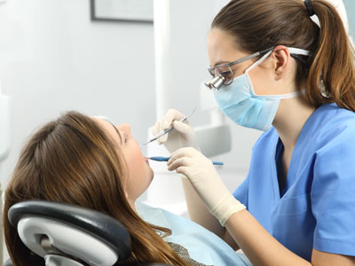 A dental hygienist in a white coat and mask is performing a teeth cleaning procedure on a seated woman with a smile, both are in a dental office setting.