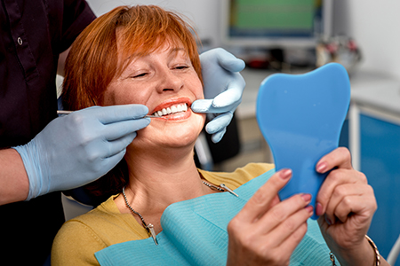 A woman with red hair sitting in a dental chair, smiling at the camera while holding up a blue model tooth.