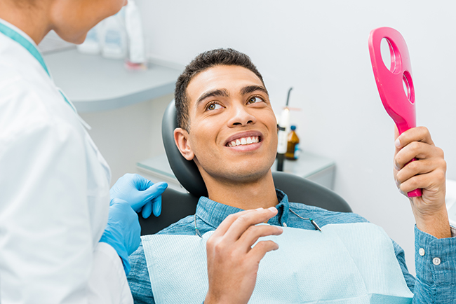 The image shows a person sitting in a dental chair with a smile, holding an oral hygiene device, while being attended to by a dental professional who appears to be providing care.