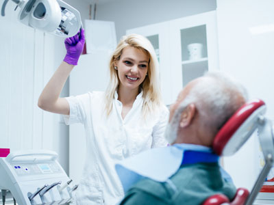 A woman wearing a white coat and purple gloves stands next to an older man sitting in a dental chair while being attended to by a dentist, with various dental equipment visible around them.