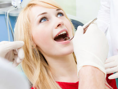 The image shows a young woman seated in a dental chair with her mouth open, receiving dental care from a professional using various tools visible around her face.