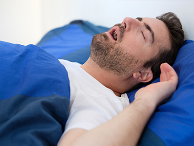 The image shows a man lying in bed with his eyes closed, appearing to be asleep, with a beard and dark hair. He has a pillow under his head and is wearing a white t-shirt. The room has blue walls and a blue blanket on the bed.